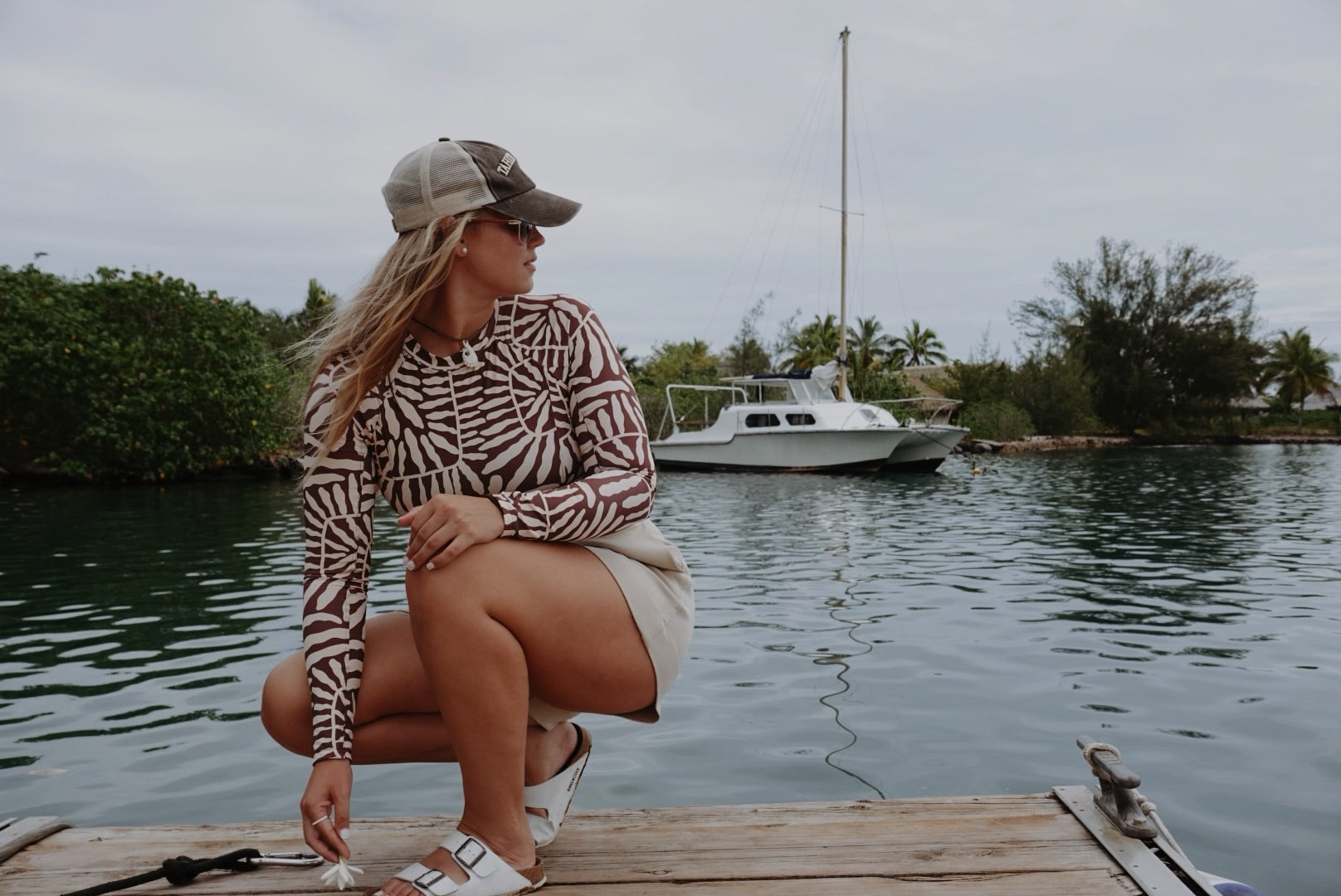A woman crouching on a wooden dock by the water, wearing a brown and beige zebra-print long-sleeve top, a beige baseball cap, and white shorts, with a boat and green shoreline in the background.