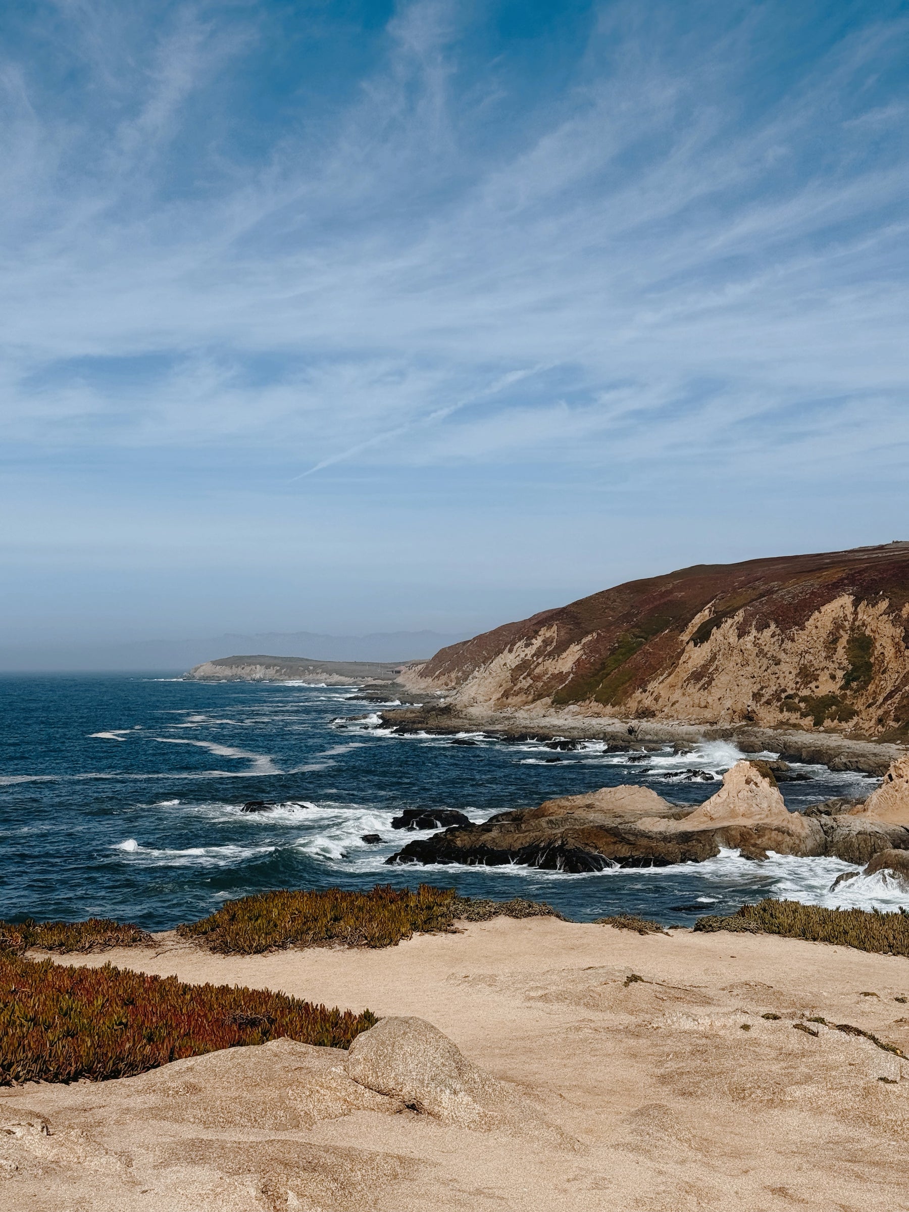 Coastal landscape with blue ocean, rocky shoreline, sandy foreground, and a brown hillside under a pale blue sky with wispy white clouds.