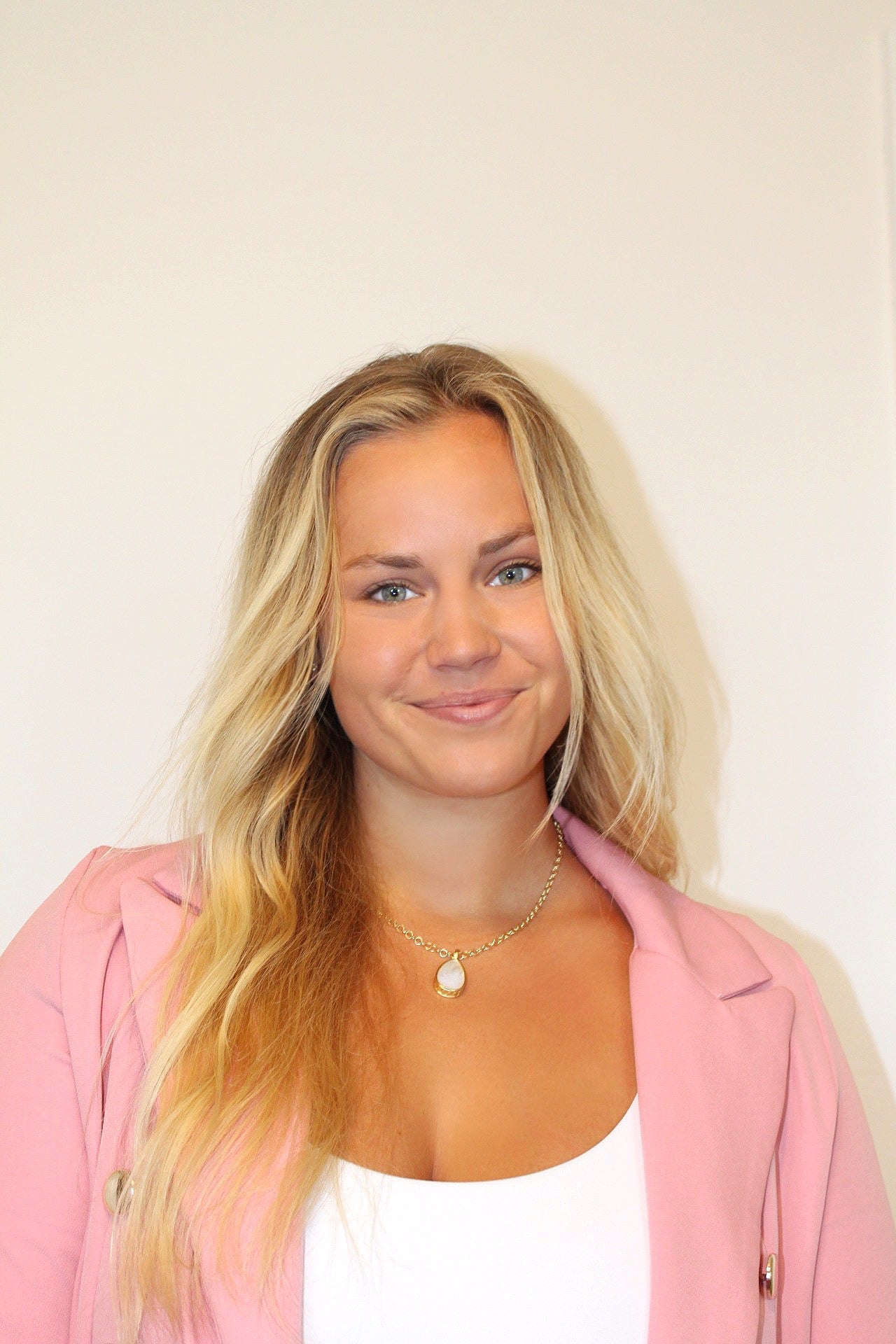 A woman with long blonde hair wearing a pink blazer over a white top, accessorized with a delicate gold necklace, photographed against a plain off-white background.