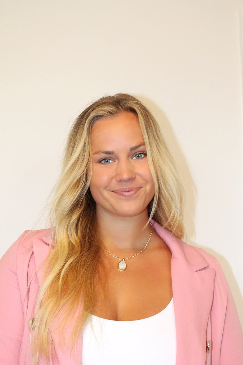 A woman with long blonde hair wearing a pink blazer over a white top, accessorized with a delicate gold necklace, photographed against a plain off-white background.