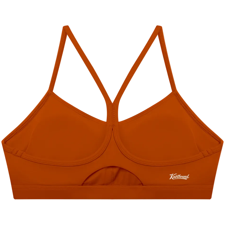 A rust-orange sports bra with a crossover criss-cross back strap design and a small white script logo on the lower right side, displayed against a solid white background.