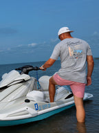 A man wearing a grey stonewash Knotbound graphic t-shirt, pink shorts, and a white cap, standing in shallow water beside a jet ski.