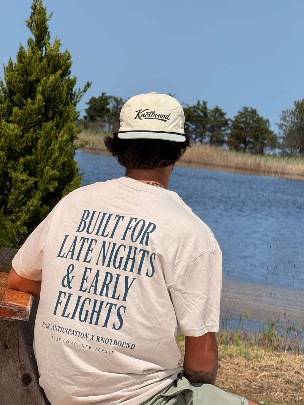A person sitting outdoors by a lake wearing a cream T-shirt with large navy text on the back and a cream Knotbound cap, photographed from behind.