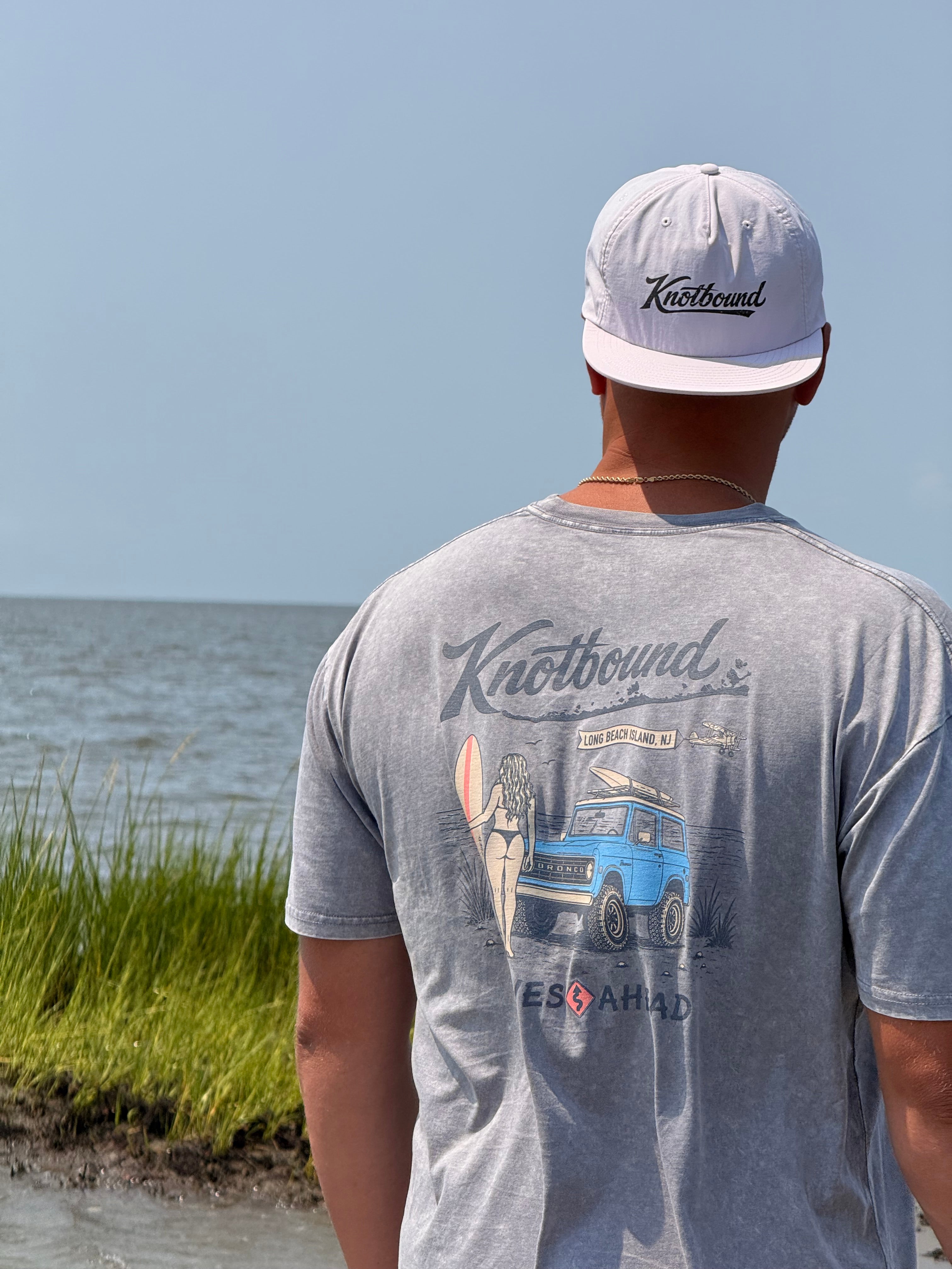 A man standing outdoors by the water, seen from behind, wearing a heather grey graphic tee and a Knotbound Surf Cap.