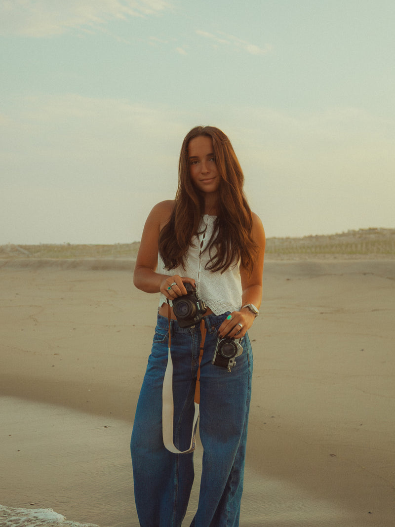 A woman standing on a sandy beach holding vintage cameras, wearing a white sleeveless top and wide-leg blue jeans.