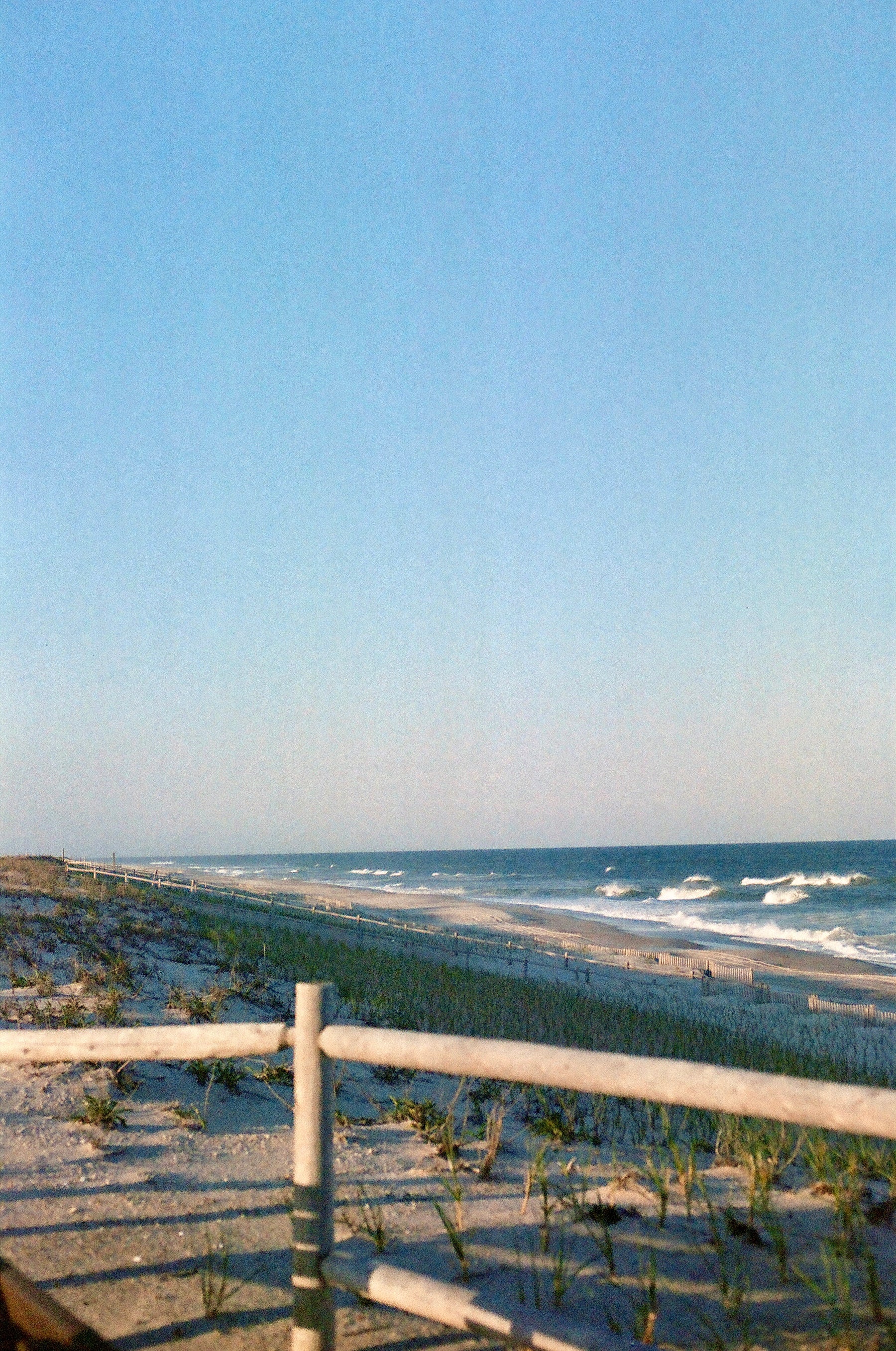 A scenic beach landscape with dunes, a wooden railing in the foreground, ocean waves, and a clear blue sky.