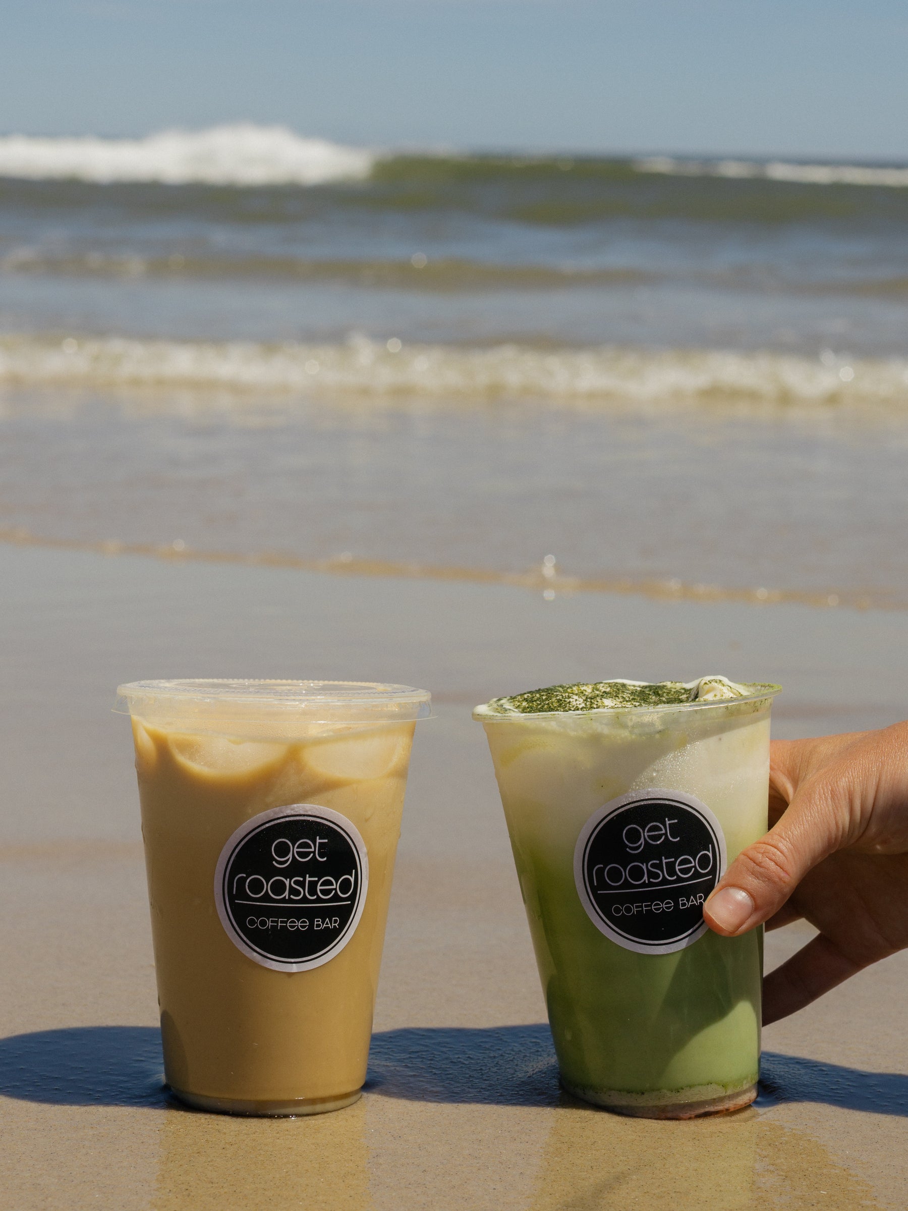 Two plastic cups of iced coffee from Get Roasted Coffee Bar sit on wet sand with the ocean waves in the background. One cup contains a tan coffee drink and the other contains a green matcha coffee drink, with a hand holding the matcha cup.