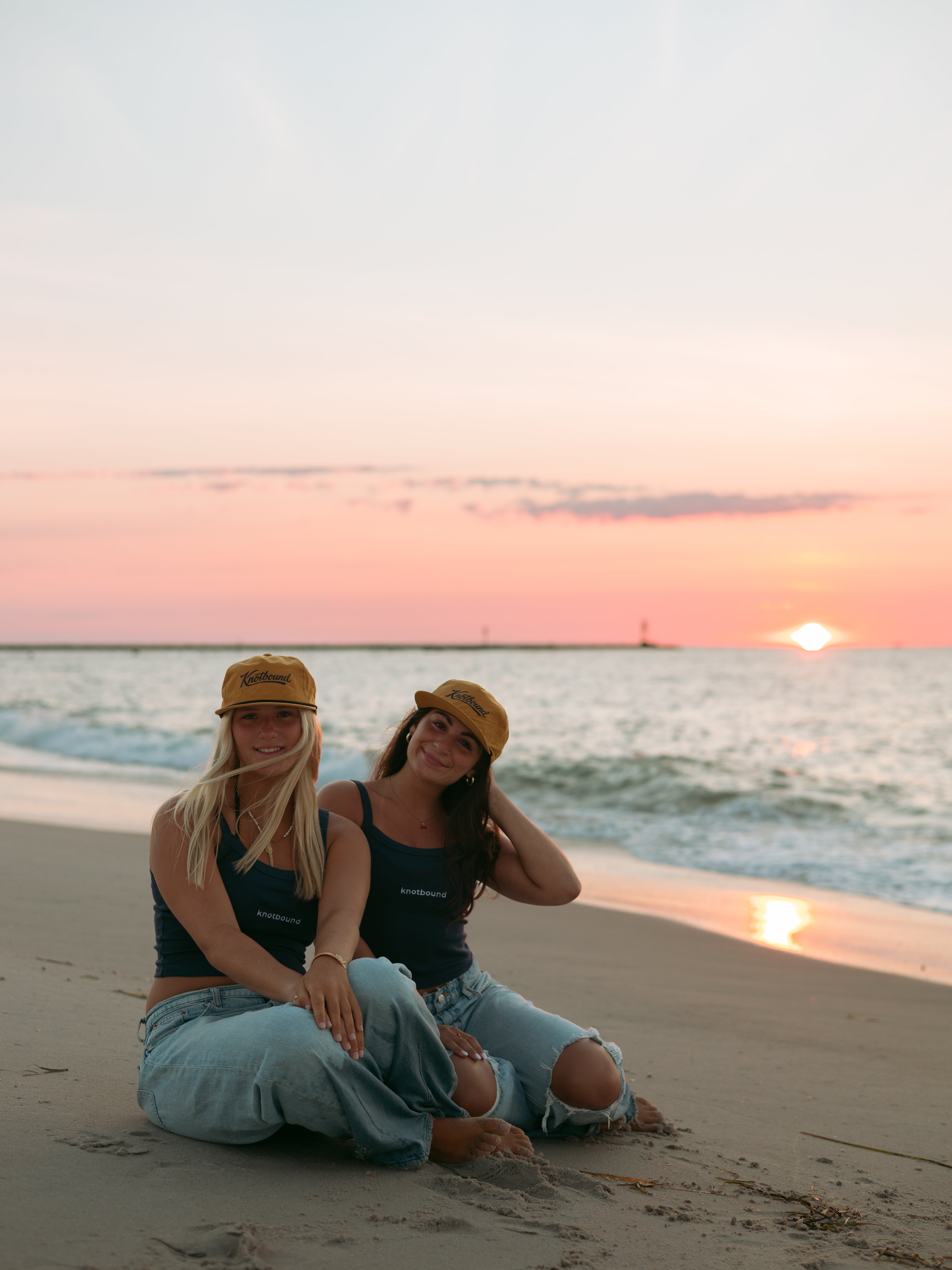 Two women sitting on a sandy beach at sunset, wearing matching navy blue tank tops, ripped light-wash jeans, and tan baseball caps.
