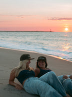 Two women sitting on a sandy beach at sunset, wearing matching dark navy blue tank tops and baseball caps, with the ocean and warm pink-orange sky behind them.