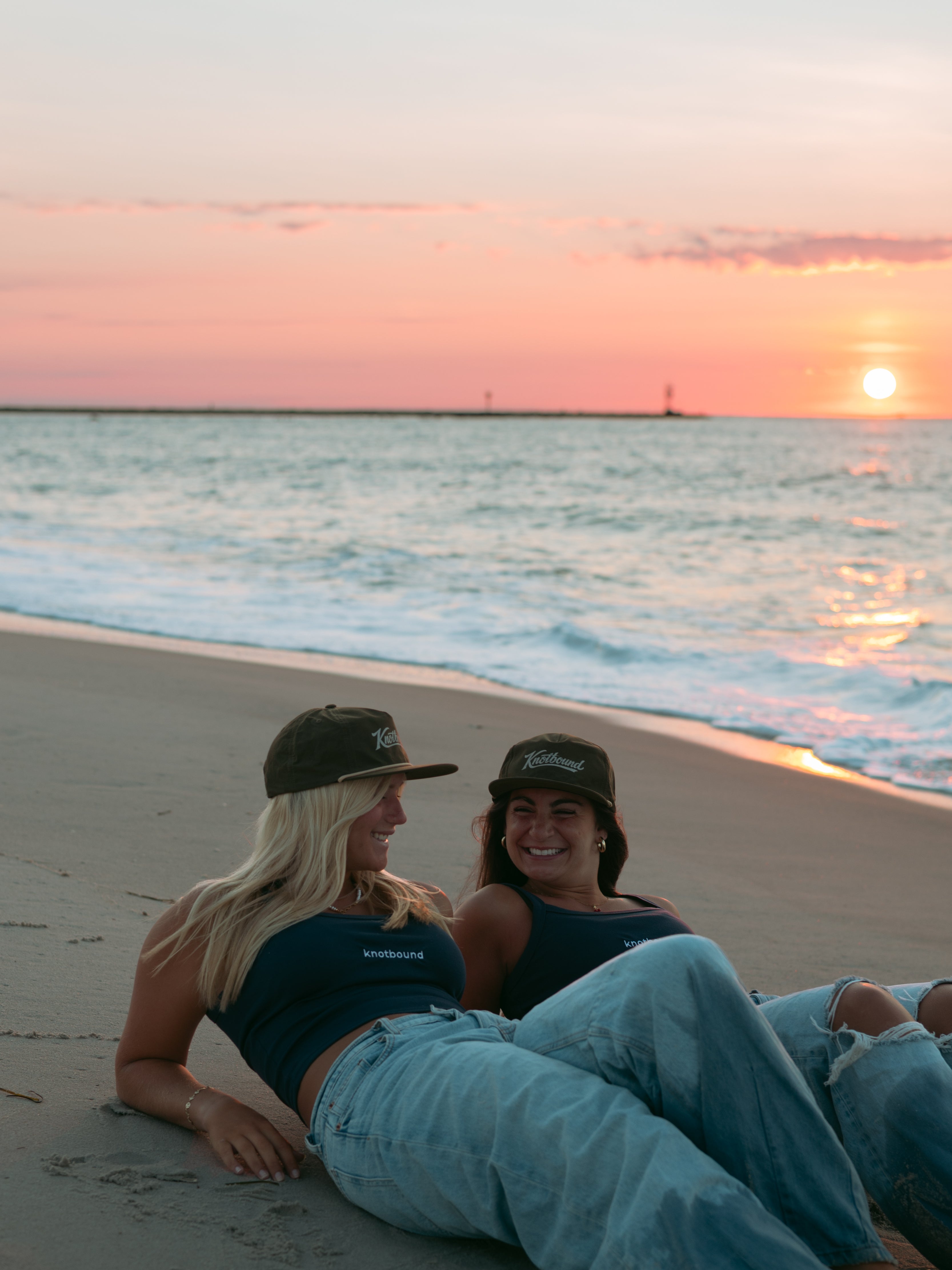 Two women sitting on a sandy beach at sunset, wearing matching dark navy blue tank tops and baseball caps, with the ocean and warm pink-orange sky behind them.