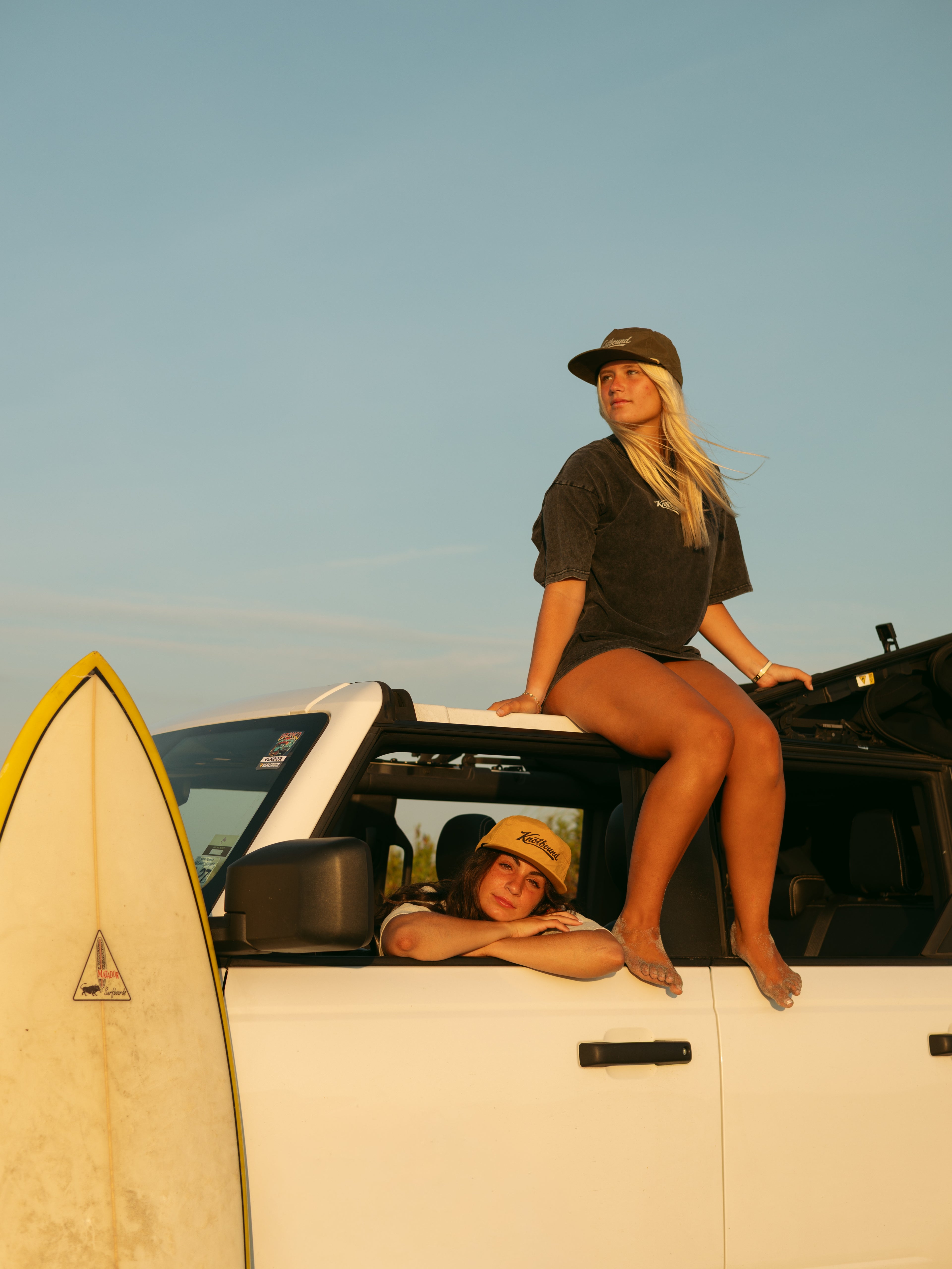 Two women posing on and inside a white vehicle outdoors under a clear blue sky, with a surfboard visible on the left. One woman sits on the roof window wearing a dark t-shirt and shorts, and the other leans out of the driver's side window wearing a tan cap.