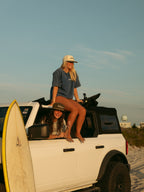 A woman in a blue t-shirt and beige shorts sits on the roof of a white off-road truck on a beach, with a yellow surfboard leaning against the front of the vehicle. Another woman is visible inside the truck.