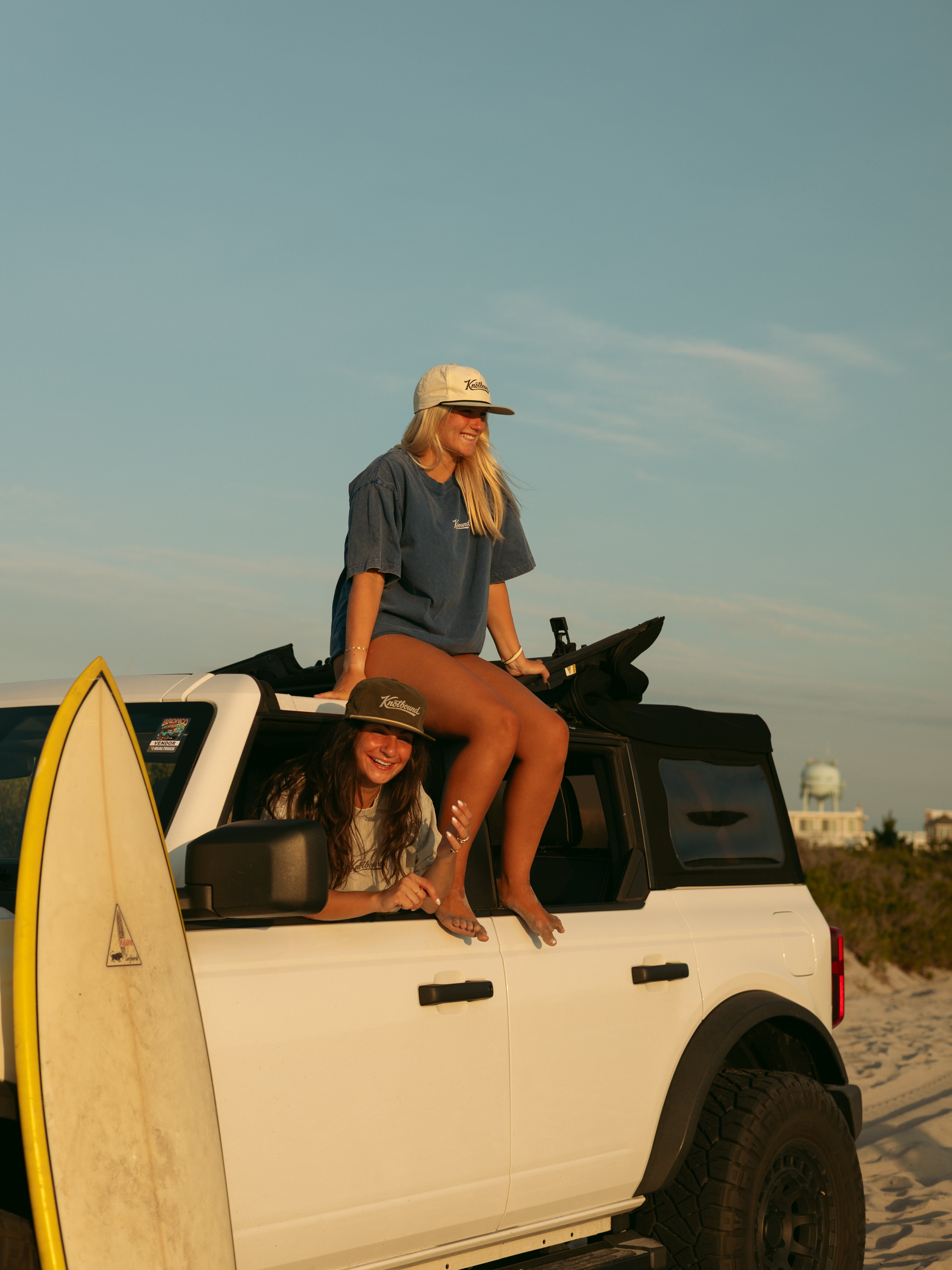 A woman in a blue t-shirt and beige shorts sits on the roof of a white off-road truck on a beach, with a yellow surfboard leaning against the front of the vehicle. Another woman is visible inside the truck.