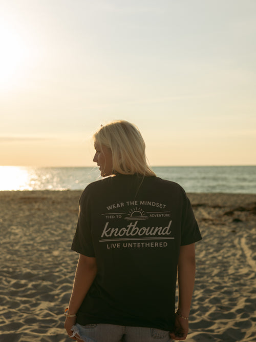 A woman on a sandy beach at sunset facing the ocean, wearing the black Knotbound Departure Tee with its Live Untethered back graphic.